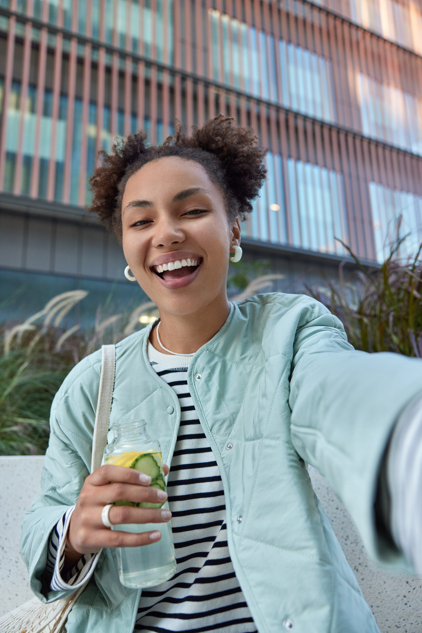 Happy optimistic girl with two hair buns dressed in jacket enjoys free time and walking in city holds bottle of detox drink makes selfie poses against modern building has fun during daytime.